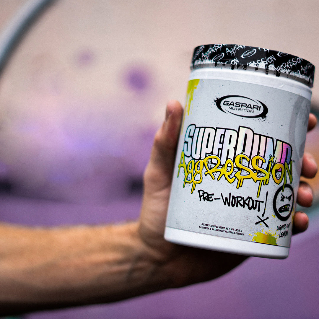 A hand holding a container of SuperPump Aggression pre-workout against a blurred gym backdrop, capturing a moment of fitness motivation.